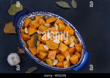 Kürbisscheiben mit Knoblauch werden in eine keramische Form auf dunklem Hintergrund, Platz kopieren, horizontale Ausrichtung, Nahaufnahme Stockfoto