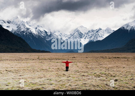 Mann im roten Mantel Stand hielt seine Arme mit Blick auf die schneebedeckten Berge, Fiordland Nationalpark, UNESCO, South Island, Neuseeland, Pazifische Stockfoto