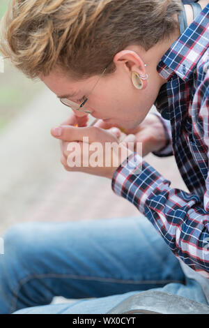 Bild vertikal an einen entspannten blonde Junge tragen ein Quadrat shirt Beleuchtung Zigarette mit den Hintergrund unscharf. Stockfoto