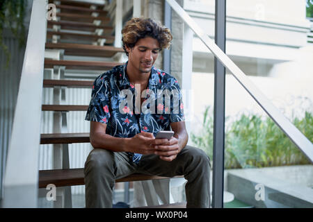 Attraktive hispanic Hipster tausendjährigen Blogger am Telefon sitzen auf moderne Treppe der helle Zimmer mit tropischen Pflanzen suchen Stockfoto