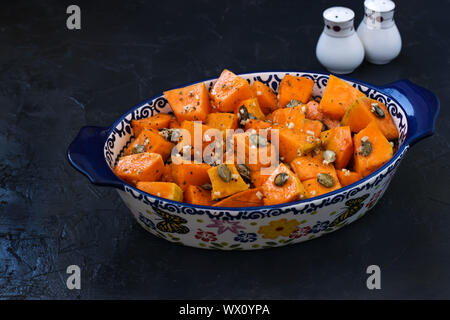 Vorbereitung für das Backen Kürbisscheiben mit Knoblauch und Kürbiskerne in eine keramische Form auf einem dunklen Hintergrund, horizontale Ausrichtung, Nahaufnahme Stockfoto