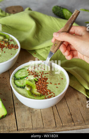 Diätetische Smoothies mit Avocado, Gurke, Kiwi und Leinsamen in eine weiße Platte auf einem Holzbrett. Hand der Frau hält einen Löffel Stockfoto