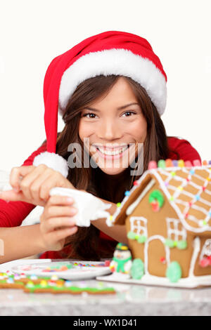 Weihnachten Mädchen, Gingerbread House. Junge Frau in Santa Hut tun Weihnachtsvorbereitungen, Sahnehäubchen Lebkuchenhaus. Gemischte Rasse/Asiatisch Stockfoto