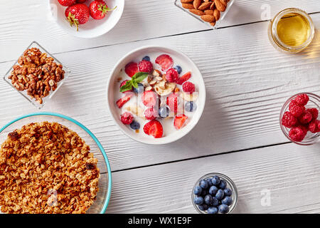 Hausgemachtes Müsli in einer Platte, Bananenscheiben, Beeren, Mandeln, Walnüsse und weiße Schüssel mit natürlichen organischen Frühstück auf weißen Woo Stockfoto