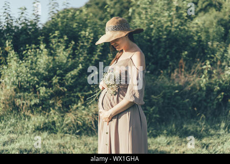 Schwangere Frau in kleid und hut außerhalb mit einem Blumenstrauß Stockfoto