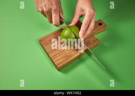 Hände einer Frau eine natürliche Grün reif Kalk in der Hälfte auf einer hölzernen Schneidebrett auf grünem Hintergrund mit Kopie Raum. Stockfoto