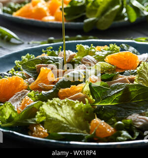 Ein Strom von Olivenöl gießt in die vorbereiteten Salat mit natürlichen, grünen Gemüse, Huhn, orange Scheiben in einem blauen Ceram Stockfoto