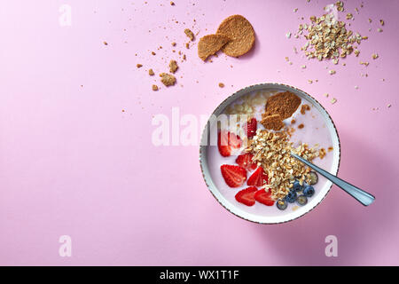 Eine weiße Schale mit organischen Joghurt Smoothie mit Erdbeeren, Banane, Blaubeere, Haferflocken und Kekse auf rosa Hintergrund. Flat l Stockfoto