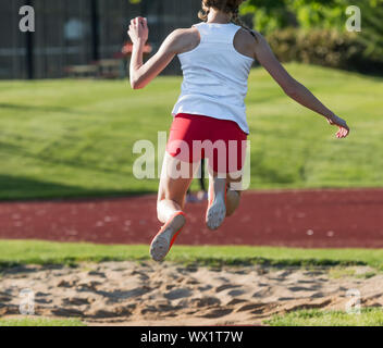 Ansicht von hinten von einem Jugendmädchen triple an einem Leichtathletik Wettbewerb springen, fliegen in die Luft Richtung Sandkasten. Stockfoto