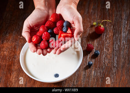 Womans Hände Vorbereitung gesunde natürliche Frühstück fron reife Beeren und organische Milch in einer Schüssel auf einem Holztisch. Stockfoto