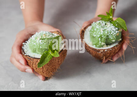 Ein Mädchen halten Sie die beiden Hälften der Kokosnuss mit grüner Minze Sorbet mit Zweig Minze und Kokos Chips auf grauem Beton. Stockfoto