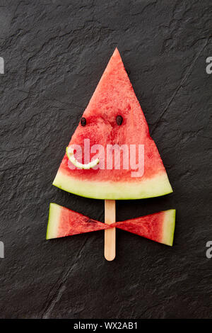Ein saftiges Stück Wassermelone auf einem Stock in Form von einem lächelnden Gesicht auf einem dunklen Schiefer Hintergrund mit kopieren. Flach Stockfoto