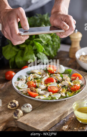 Mans Hand ein Foto von frisch zubereiteten hausgemachten Salat mit natürlichen Zutaten von Smartphone über eine hölzerne Hintergrund. Stockfoto