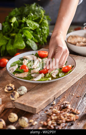 Der Frau die Hand ein Stück Tomate in einem Teller mit Spinat und Fleisch auf einem Holztisch. Salat Vorbereitung. Stockfoto