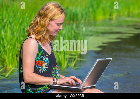 Junge Frau arbeitet auf dem Laptop in der Nähe von Wasser in der Natur Stockfoto