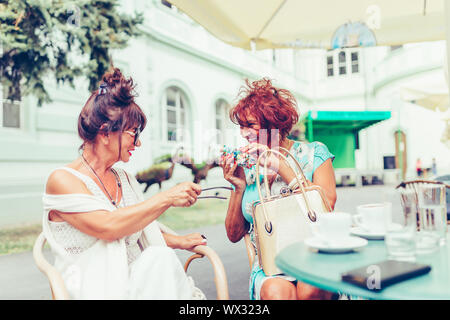 Zwei glückliche älterer Frauen sprechen und trinken Kaffee sitzen, die in der Tabelle in einem Café im Freien. Ältere Frau, ihren Freund, ihre neue Brille angezeigt. Stockfoto