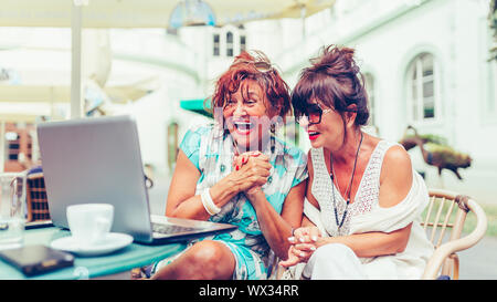 Zwei aufgeregt älterer Frauen lesen gute Nachrichten online auf Laptop beim Sitzen in einem Café im Freien. Stockfoto