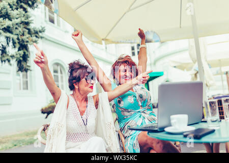 Zwei aufgeregt älterer Frauen lesen gute Nachrichten auf der Linie mit einem Laptop im Café im Freien. Stockfoto