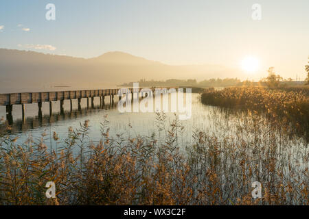 Lange Holzsteg pier über Wasser in goldenen Abendlicht mit einer Berglandschaft silhouette i Stockfoto