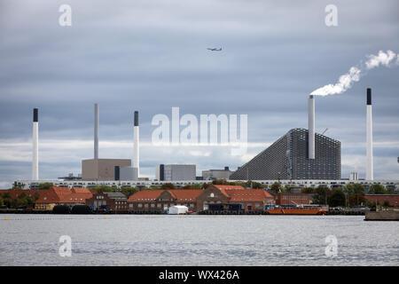 Ein Flugzeug fliegt über den Amager bakke Kombiniert tnv Anlage und Skipiste in Kopenhagen, Dänemark, mit dem Hafen im Vordergrund. Stockfoto