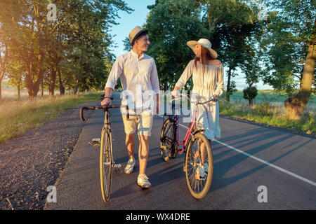Süßes Paar wheeling Fahrräder und fröhlich plaudernd auf einer Sommer Landstraße durch Sonnenuntergang Sonne beleuchtet Stockfoto