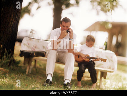 Ein Junge klimpert seine Gitarre, während sein Vater auf einem Mobiltelefon Gespräche. Stockfoto