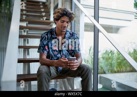 Attraktive hispanic Hipster tausendjährigen Blogger am Telefon sitzen auf moderne Treppe der helle Zimmer mit tropischen Pflanzen suchen Stockfoto