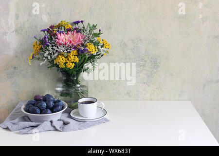 Feminine Hochzeit, Geburtstag Szene mit Tasse Kaffee und Pflaume Obstschale. Glas Vase mit schönen bunten Strauß von Dahlia, tansies und Aster flowe Stockfoto