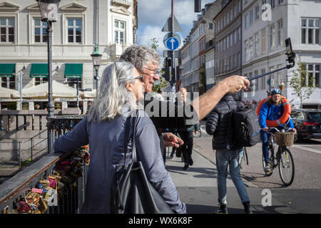 Ein Paar mit einem selfie-Stick ein eigenes Bild auf dem Nyhavnsbroen Brücke zu nehmen, Nyhavn, in Kopenhagen, als Radfahrer mit einem rettungsring Fahrten Vergangenheit Stockfoto