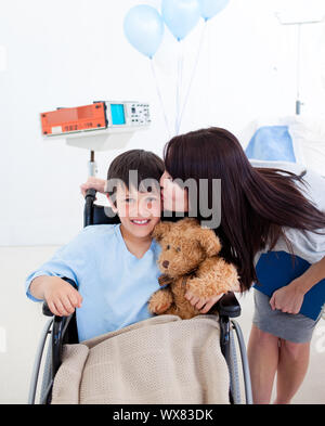 Fröhlich lächelnd wenig Junge sitzt im Rollstuhl und seine Mutter im Krankenhaus Stockfoto