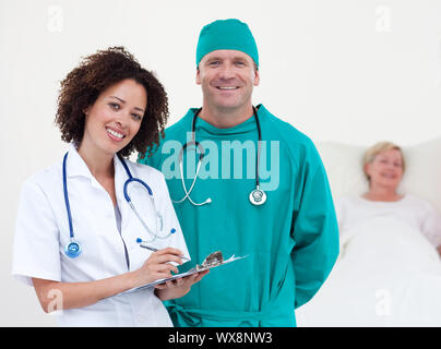 Charismatische Team von Ärzten an Kamera liegenden Patienten in ihrem Bett Lächeln in einem Krankenhaus Stockfoto