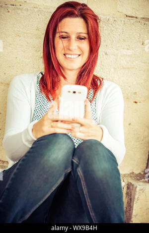 Hübsches Lächeln rothaarige Frau gegen eine alte Mauer in den Prozess der Übermittlung von Nachrichten per Handy sitzen Stockfoto