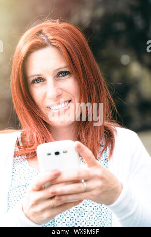 Hübsches Lächeln rothaarige Frau, die das Senden von Nachrichten oder ein selfie auf Ihr Handy machen, gegen den Hintergrund der Sommer Green Park. Stockfoto