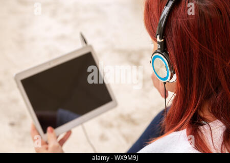 Hübsches Lächeln rothaarige Frau auf dem Boden sitzend hören von Musik oder beim Anschauen eines Films auf einem Touch Pad. Fokus auf Kopfhörer Stockfoto