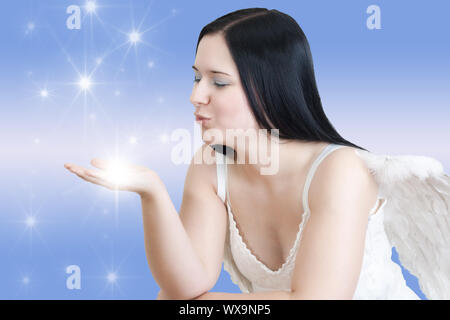 Junge schwarzhaarige Frau mit Engelsflügeln weht einige Sterne auf blauem Hintergrund Stockfoto