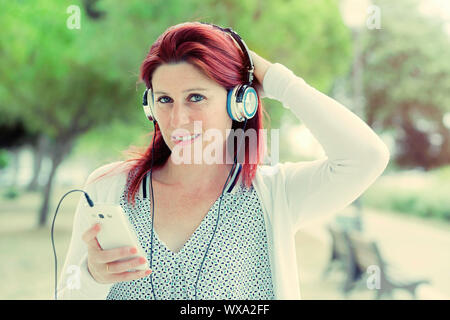 Attraktive lächelnd rothaarige Frau Hören von Musik auf Ihrem Handy mit dem Grün im Hintergrund Stockfoto
