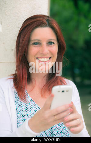 Hübsches Lächeln rothaarige Frau Senden von Nachrichten auf Ihr Handy in einem Park Stockfoto