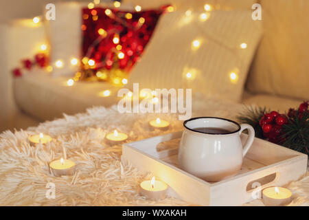 Heiße Tasse Tee in einem weißen Becher, Kerzen und Lichterketten auf einem Sofa mit Kissen, modernen Interieur Stockfoto