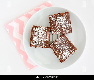 Hausgemachte Schokolade brownies Quadrate mit Abstauben des Puderzucker auf weiße Platte mit rosa Serviette. Auf weißem Hintergrund im Querformat in Flach Stockfoto