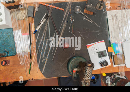 Blick von oben auf den Arbeitsplatz einer Kunst Glasbläser mit Gasbrenner, diverse Utensilien und unvollendete Artefakte in einem glasbläser Manufaktur. Stockfoto