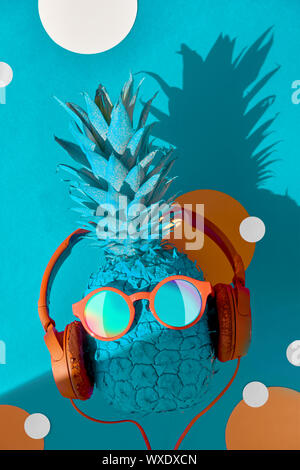 Lustige Ananas in Sonnenbrille und Kopfhörer mit einem Schatten, flach auf blauem Papier Hintergrund Stockfoto