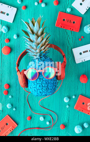 Lustige Ananas in Sonnenbrille und Kopfhörer blau lackiert. Flach auf rotem Papier Hintergrund mit weichen Bällen und retro Tonbänder. Stockfoto