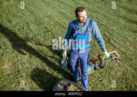 Porträt eines professionellen Gärtner in Arbeitsschutzausrüstungen ruht beim Schneiden von Gras mit Benzin Rasenmäher auf dem Hinterhof, Ansicht von oben Stockfoto