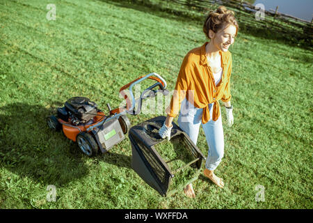 Schöne junge Frau mit Korb voller Gras bei der Gartenarbeit mit Rasenmäher auf dem Hinterhof in der Landschaft Stockfoto