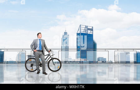 Junger Mann mit Anzug stehen auf Balkon mit dem Fahrrad. Geschäftsmann mit dem Fahrrad auf dem Hintergrund des blauen Himmels über Megalopolis. Männliche Radfahrer halten Stockfoto