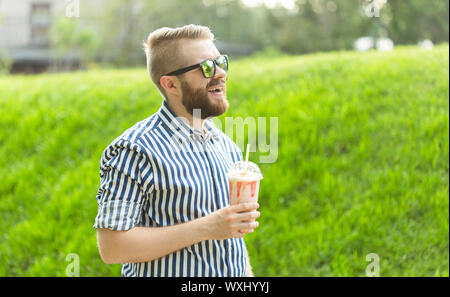 Seitenansicht eines stilvollen junger Mann mit einem Bart Holding einen Milchshake und bewundern Sie die Aussicht auf die Stadt zu Fuß in den Park an einem warmen Sommertag. Das Konzept der Stockfoto