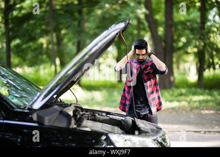 Closeup Portrait, junger Mann, haben Sie Probleme mit seinem gebrochenen Auto öffnen Motorhaube, Motor, zu beheben versuchen isoliert grüne Bäumen außerhalb Hintergrund. Auto wird nicht Stockfoto