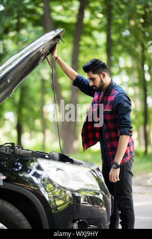 Closeup Portrait, junger Mann, haben Sie Probleme mit seinem gebrochenen Auto öffnen Motorhaube, Motor, zu beheben versuchen isoliert grüne Bäumen außerhalb Hintergrund. Auto wird nicht Stockfoto