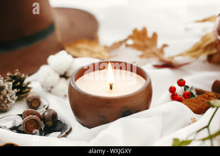 Hygge Lebensstil. Kerze, Beeren, fallen, Blätter, Kräuter, Eicheln, Nüsse und braunen Hut auf weißem Stoff. Herbstliche Stimmung. Hallo Herbst, gemütliche inspirierende Bild. Stockfoto