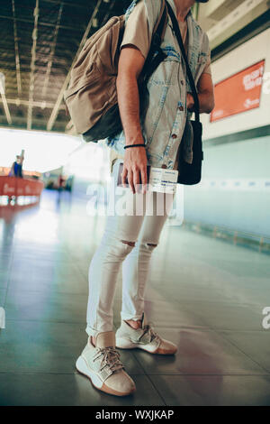 Leger gekleideter Mann mit Rucksack und Tickets unter Fahrplan ist auf der Suche beim Stehen in der Airport Terminal Hall Stockfoto